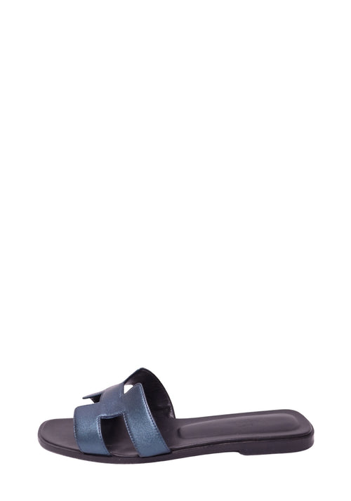 Hermès metallic blue leather flat slide sandal featuring signature H-shaped cutout strap, shimmering blue finish, open toe, dark footbed, and slim flat sole