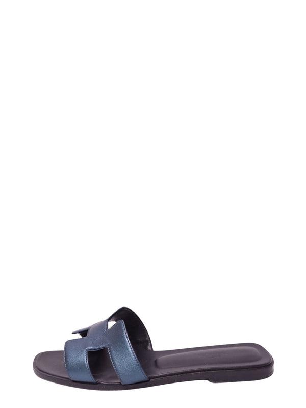 Hermès metallic blue leather flat slide sandal featuring signature H-shaped cutout strap, shimmering blue finish, open toe, dark footbed, and slim flat sole
