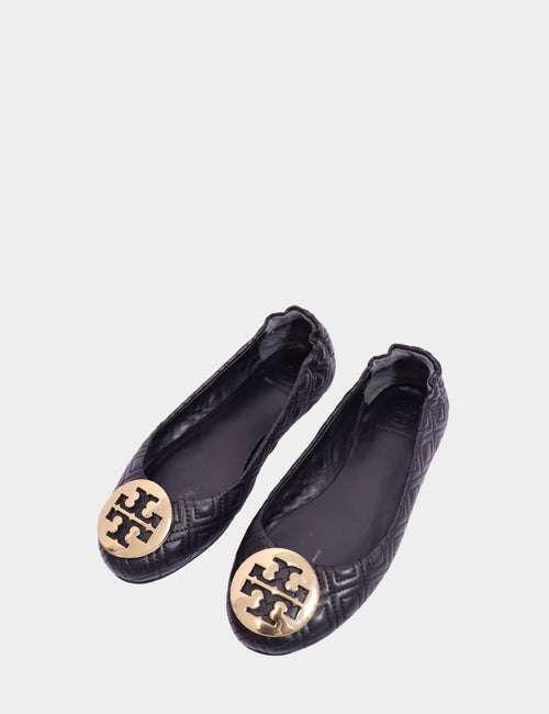 "Tory Burch black quilted ballet flats with gold logo medallion and elastic back, displayed on white background."

